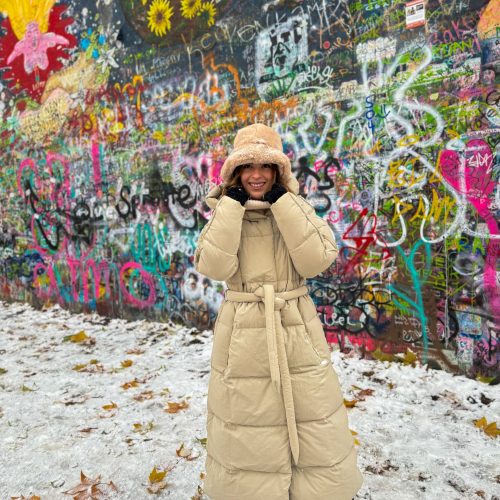 lennon-wall-anna-trakada κορίτσι με τεράστιο μακρύ μπουφάν μπροστά από τον τοίχο του Λένον στην Πράγα